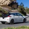 A rear side profile shot of a white 2023 Nissan Leaf all-electric hatchback driving up a curving hill road