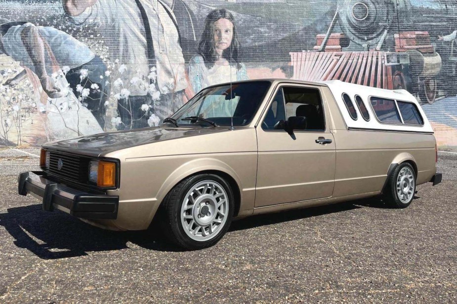 Tan VW Rabbit pickup truck with a white cap and lowered suspension is parked in front of a mural.