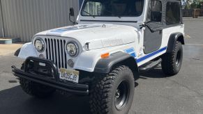 The Jeep grille of a white CJ-8 Scrambler 4x4 restomod with Mercedes turbodiesel badges, a car dealership visible in the background.