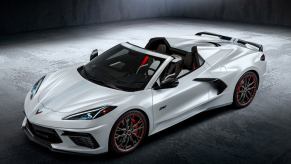 White Chevy Corvette convertible sports car parked on pavement, its crossed flags logo visible in the foreground.