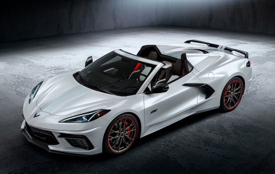 White Chevy Corvette convertible sports car parked on pavement, its crossed flags logo visible in the foreground.