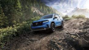 A light blue Honda Pilot driving in a wooded area on a dirt road.