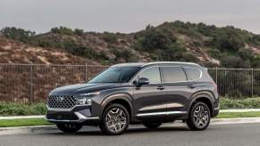 A dark grey 2023 Hyundai Santa Fe parked in front of a grassy hill on blacktop.