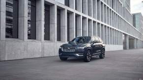 A dark color 2023 Volvo XC90 parked in front of a building lined with concrete framed windows.