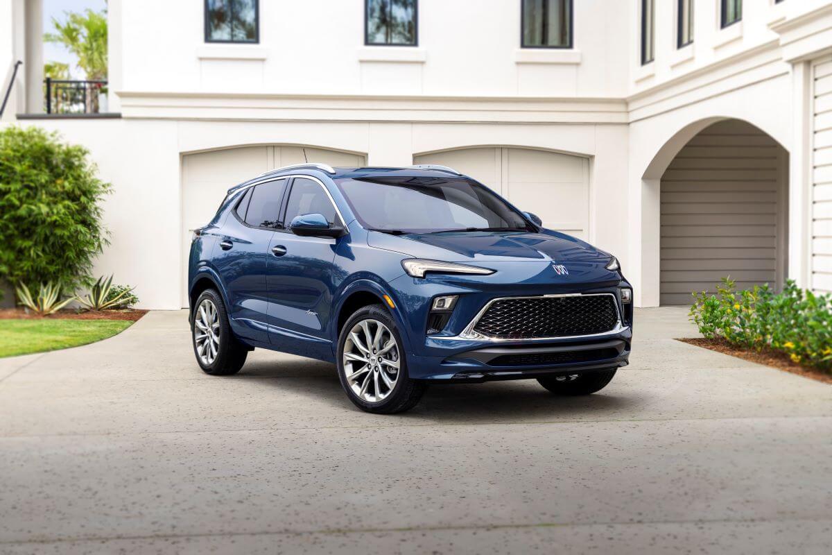 A blue 2024 Buick Encore GX Avenir subcompact crossover SUV parked on a driveway in front of a white luxury home