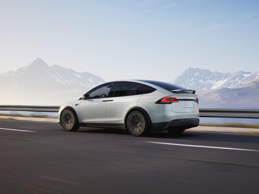 Rear view of a white Tesla Model X driving down a road with mountains in the background.