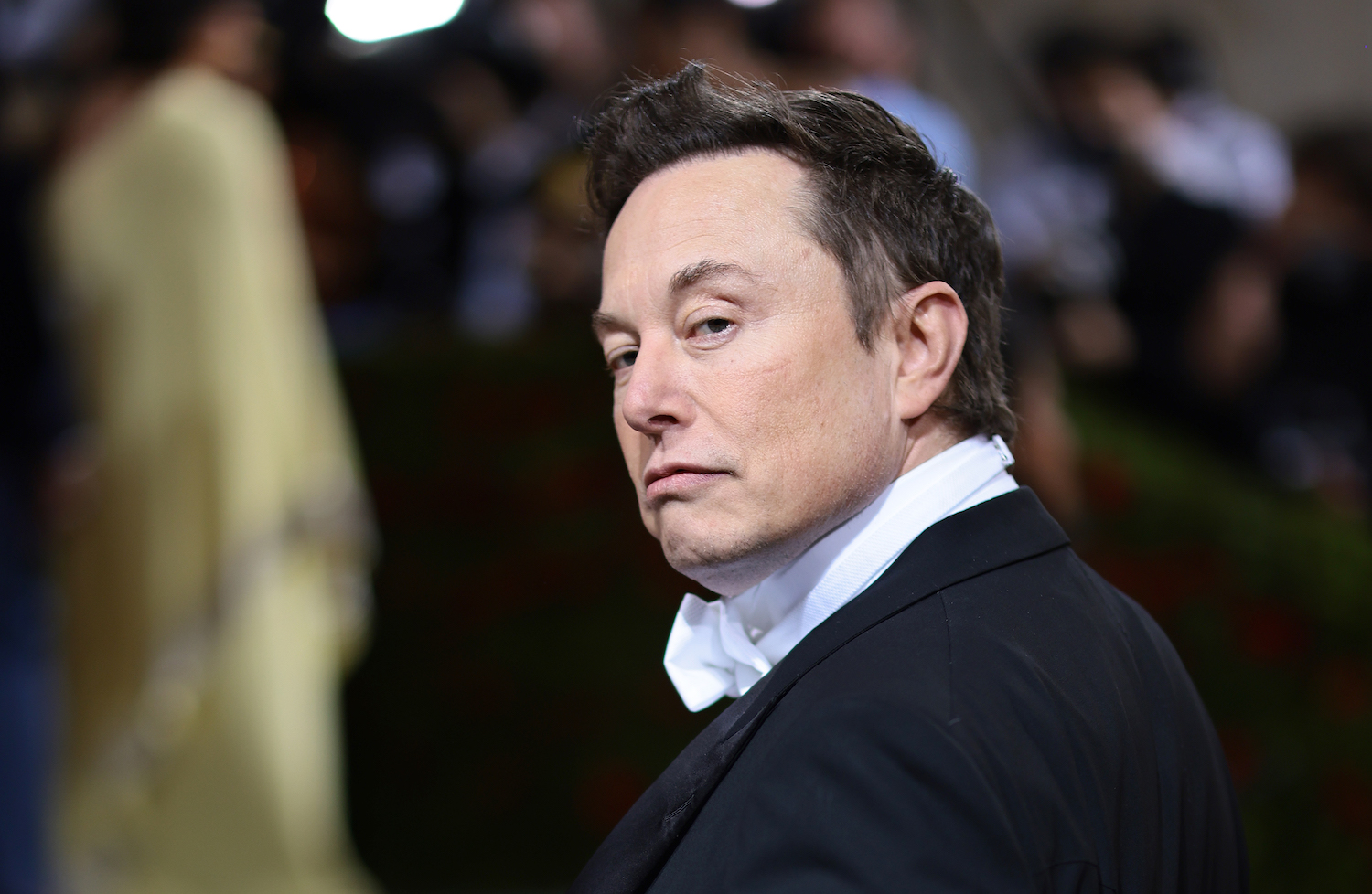 Elon Musk wearing a tuxedo at the met Gala, a blurry crowd visible behind him.