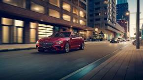 A red Genesis G70, one of the safest cars of 2022, driving down the road in a city surrounded by buildings.