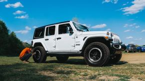 A woman crouches by a white Jeep Wrangler SUV to adjust its tire pressure, a field and blue sky visible in the background.