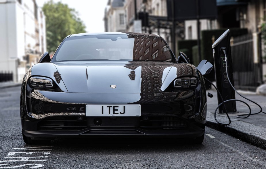 A black Porsche Taycan EV is parked on the street, connected to a charger, the building of the Mayfair district of London visible in the background.