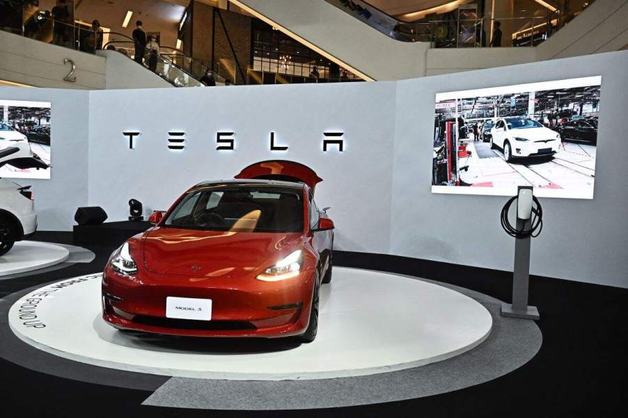 A red Tesla Model 3 on display in front of a grey wall with Tesla written on it and a screen with a video playing.