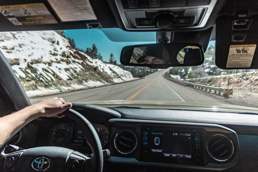 A man's hand on the steering wheel of his Toyota Tacoma pickup truck, snowy mountains visible through the windshield.