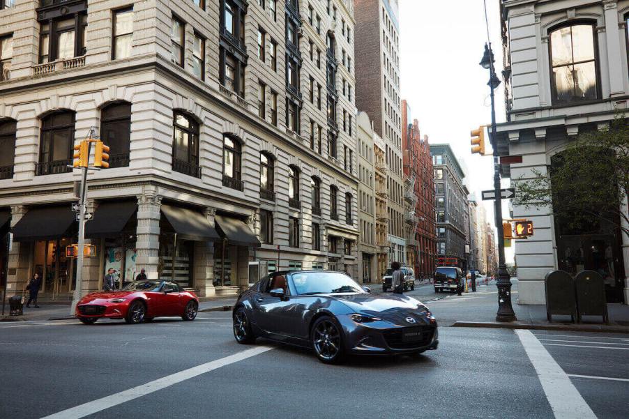 A gray Mazda MX-5 Miata turning on a city street in front of some buildings