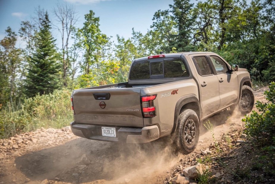 A brown Nissan Frontier Pro-4X navigates a dusty off-road trail, pine trees in the background.