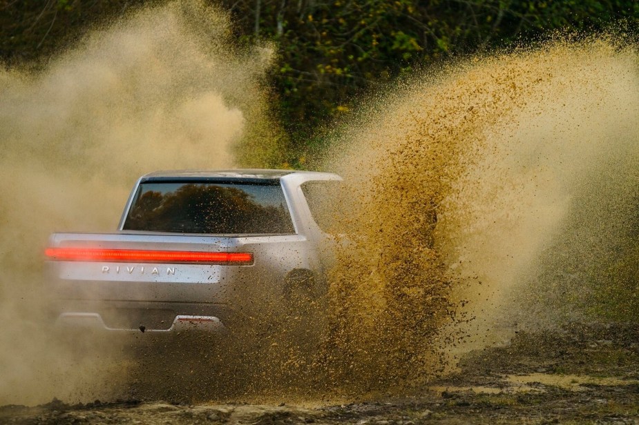 Silver Rivian R1T electric pickup truck off-roading through a muddy obstacle, dirt flying off its rear wheels.