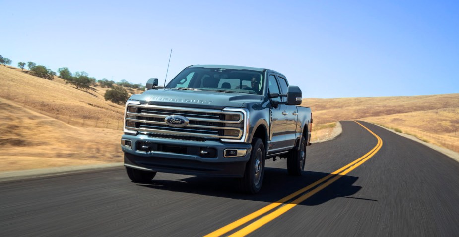 A blue Ford Super Duty truck driving down a paved road, desert ridgelines visible in the background.