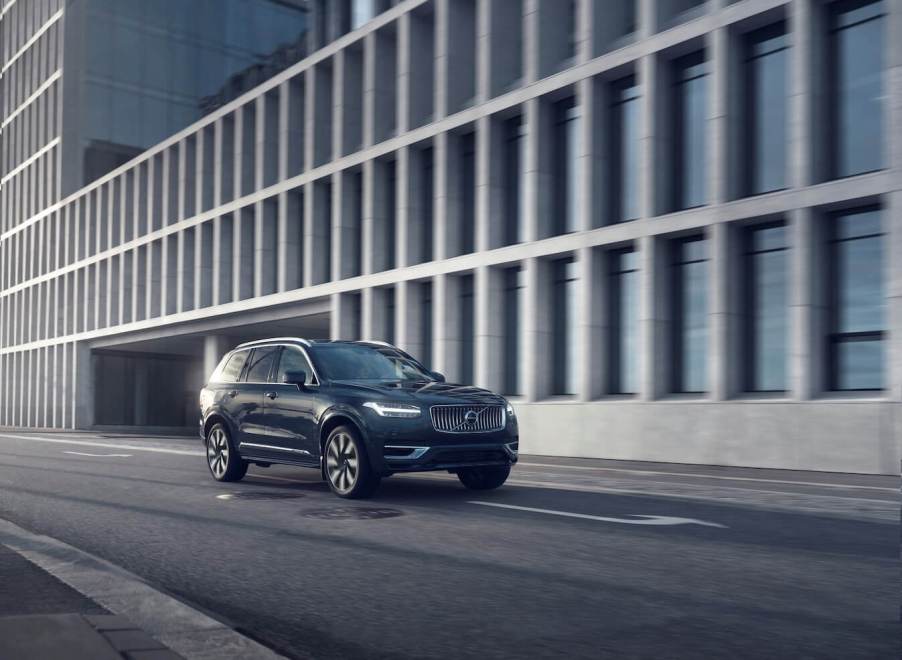 A blue 2023 Volvo XC90 Recharge driving down a city street in front of a building.