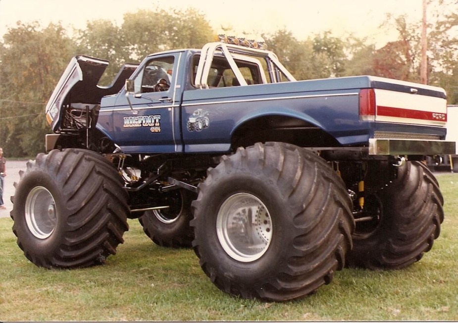 The Ford F-250 Bigfoot monster truck with its hood up and parked in a field, trees visible in the background.