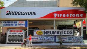 A woman pushes a stroller past a Bridgestone/Firestone tire outlet store with several vehicles on lifts having their tires changed.