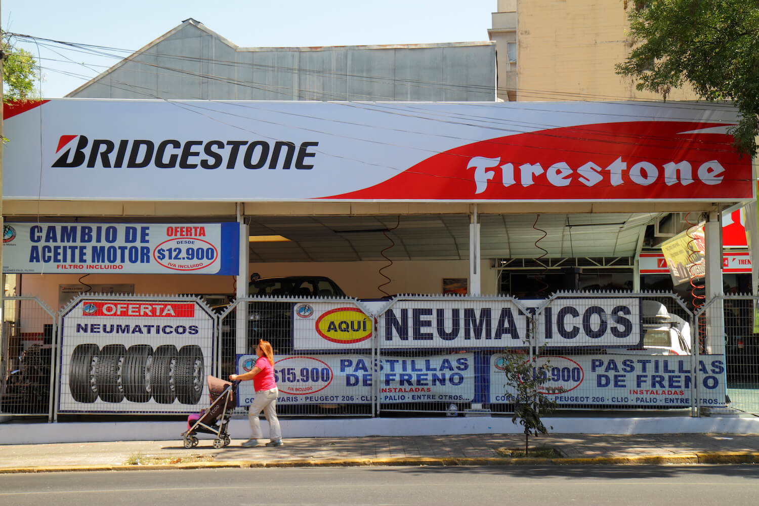 A woman pushes a stroller past a Bridgestone/Firestone tire outlet store with several vehicles on lifts having their tires changed. 