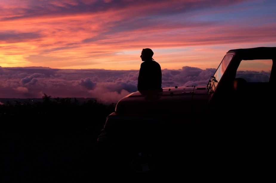 A person sits on the hood of their Jeep Wrangler as the sun sets.