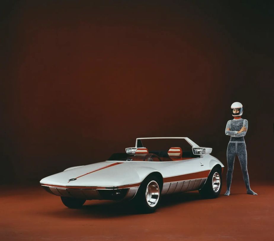 A model in a helmet and jumpsuit stands next to a 1969 Fiat prototype on stage at an auto show.