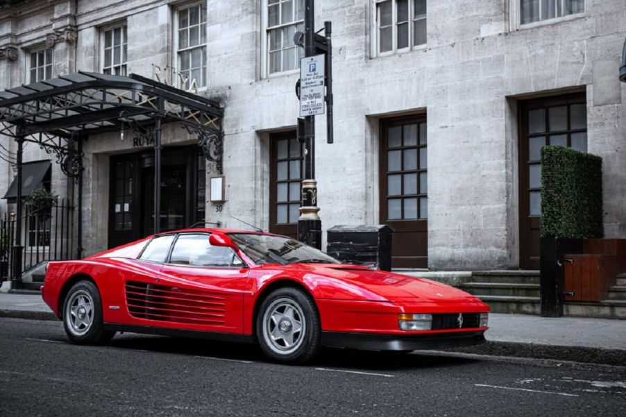 A red classic Ferrari Testarossa supercar parks next to an old building.