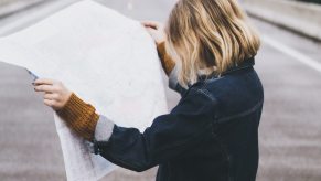 Blonde woman stands in the center of an interstate highway while examining an unfolded map.