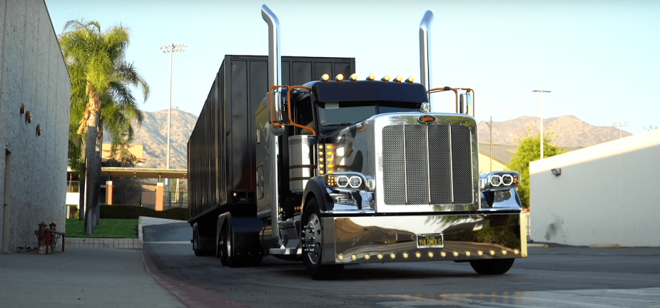 A custom Peterbilt semi-truck pulling a trailer down the road, a mountain range visible in the background.