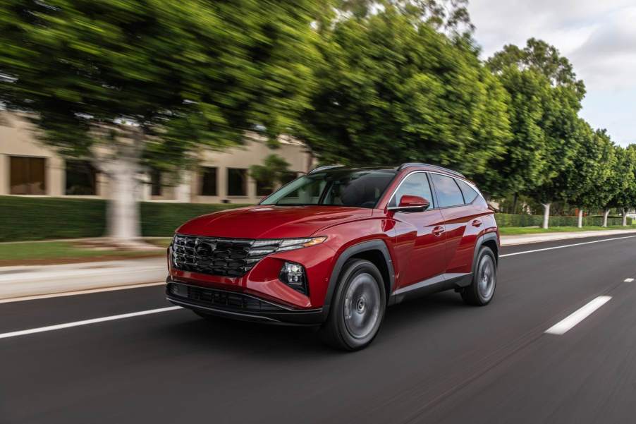 A red Hyundai Tucson drives down a city street with green trees. The Tucson is among the affordable suvs available with panoramic sunroofs.