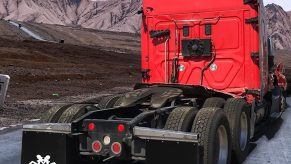 The back of a red semi-truck parked in front of a mountain range with the silhouette of a girl visible on its mudflaps.