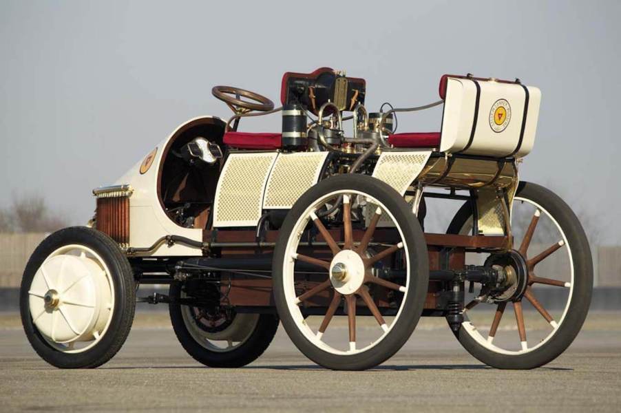 A white replica of the 1899 Porsche Semper Vivus EV parked on pavement, a fence in the background.