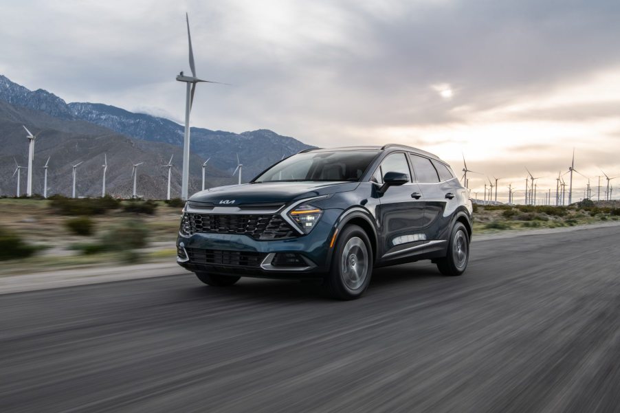 The 2023 Kia Sportage Hybrid drives in front a mountain range with a windmill in the foreground.