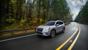 A silver Subaru Forester drivers down a wet road with trees on the side. The Subaru Forester's weakness is that it doesn't have a hybrid option.