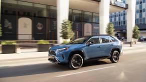 A blue Toyota RAV4 driving down a city block with business buildings in the background.