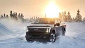 A 2022 Ford F-150 Lightning full-size pickup truck EV driving through snow as the sun sets behind evergreen trees