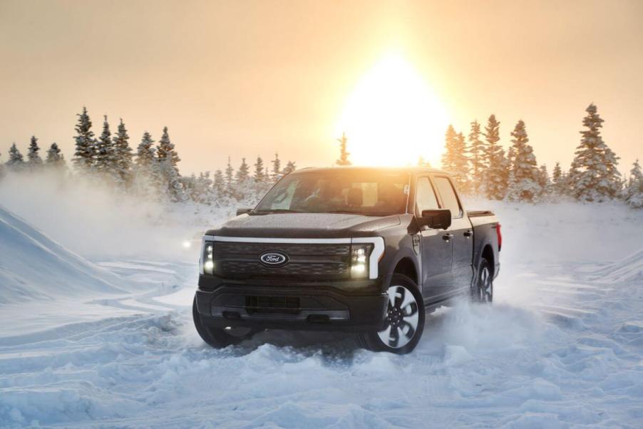 A 2022 Ford F-150 Lightning full-size pickup truck EV driving through snow as the sun sets behind evergreen trees