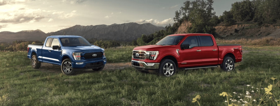 Blue and red versions of the 2022 Ford F-150, one of the least reliable 2022 pickup trucks, parked in a field of grass near a mountain range