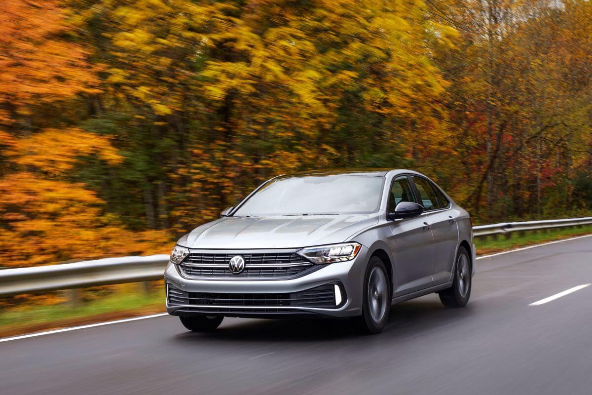 A silver-gray 2022 Volkswagen Jetta compact sedan model driving on a highway surrounded by a forest of yellow trees