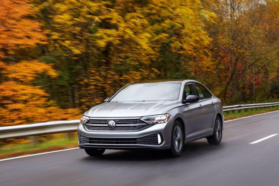 A silver-gray 2022 Volkswagen Jetta compact sedan model driving on a highway surrounded by a forest of yellow trees