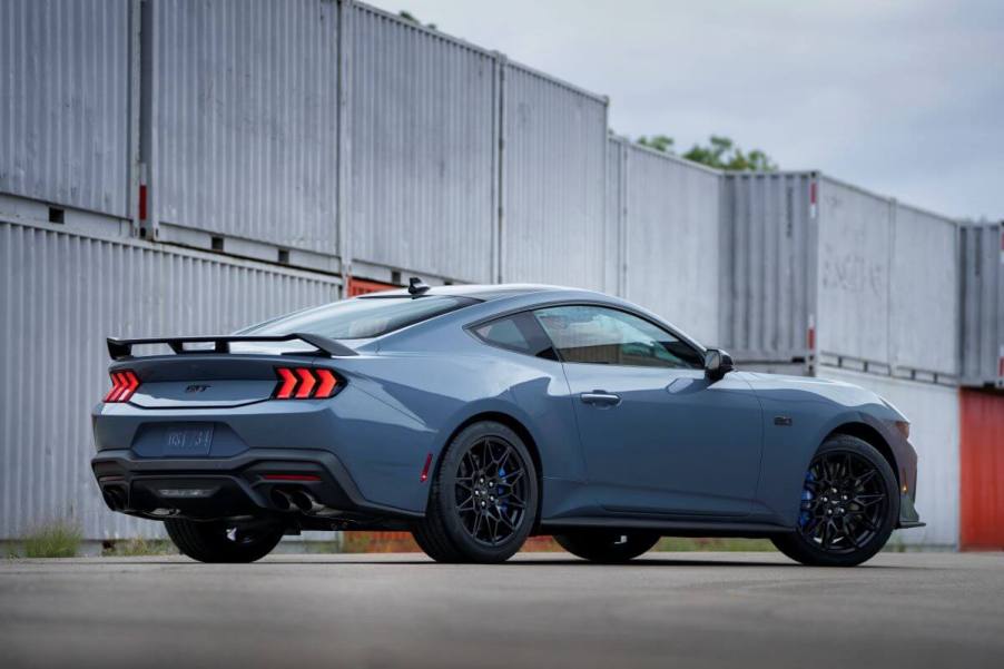 A rear exterior side shot of a blue-gray 2024 Ford Mustang muscle car coupe model parked near shipping containers. The 2024 Ford Mustang fuel economy gives it a big advantage over the competition.