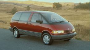 A red 1991 Toyota Previa minivan model parked on the side of a road near fields of yellow grass