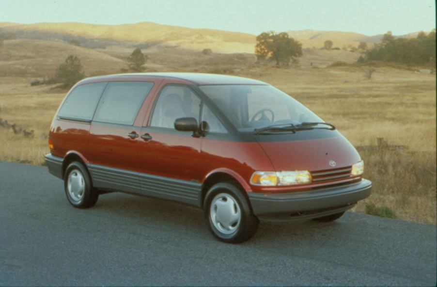 A red 1991 Toyota Previa minivan model parked on the side of a road near fields of yellow grass