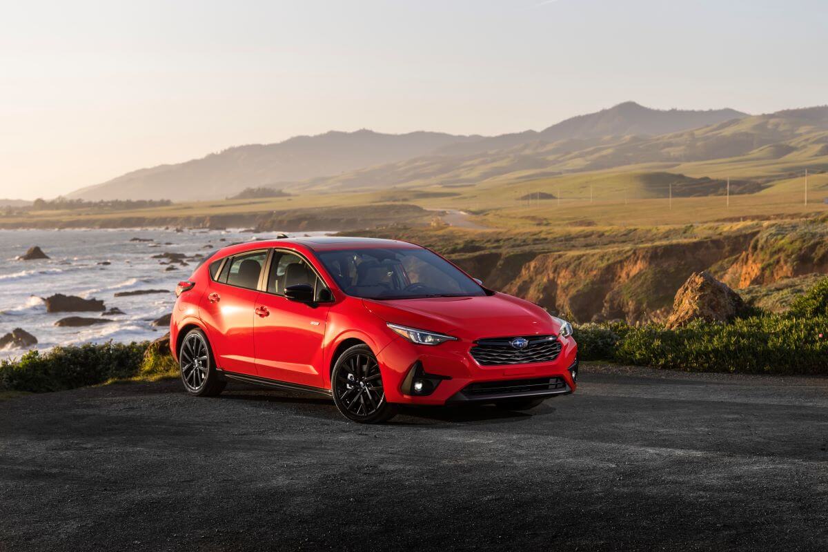 A red 2024 Subaru Impreza RS compact hatchback model parked on a grassy ledge near waves crashing in the sea