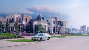 A white car parked on a city street, a big house and skyscrapers visible in the background.