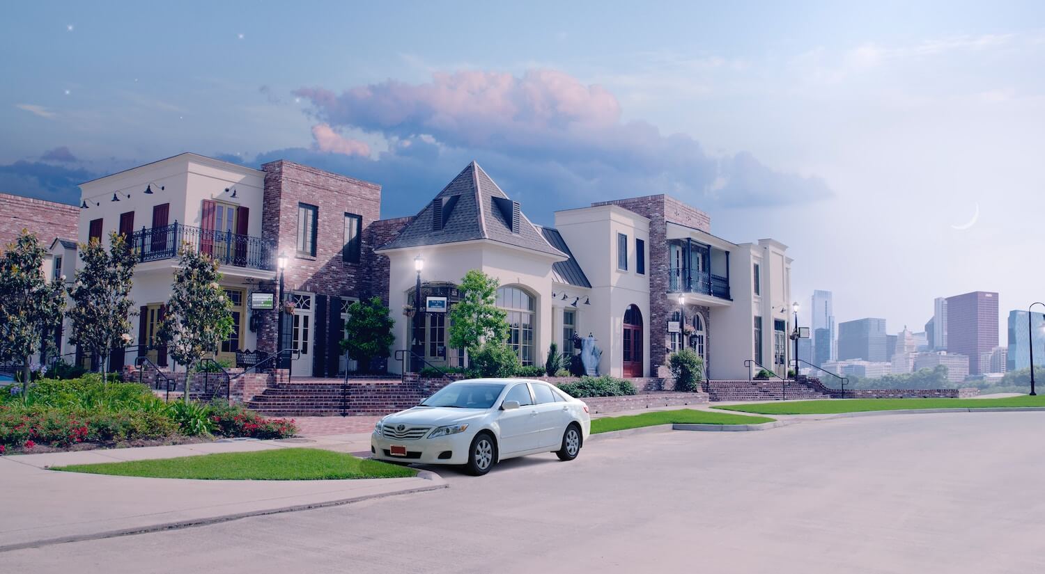 A white car parked on a city street, a big house and skyscrapers visible in the background.