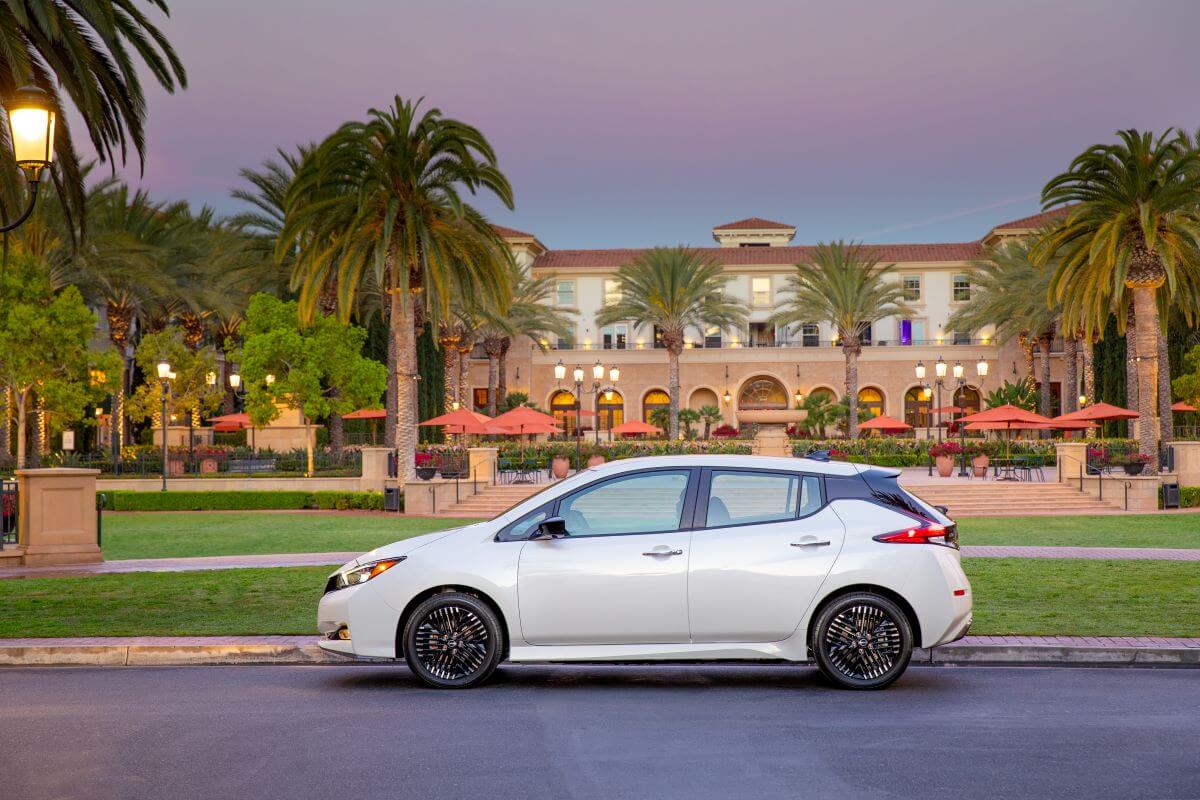 A side profile shot of a white 2023 Nissan Leaf EV hatchback model parked in front of a tropical resort