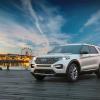 A white 2023 Ford Explorer parked on a pier with a carnival behind it. Ford sales include favorable numbers for the Explorer.