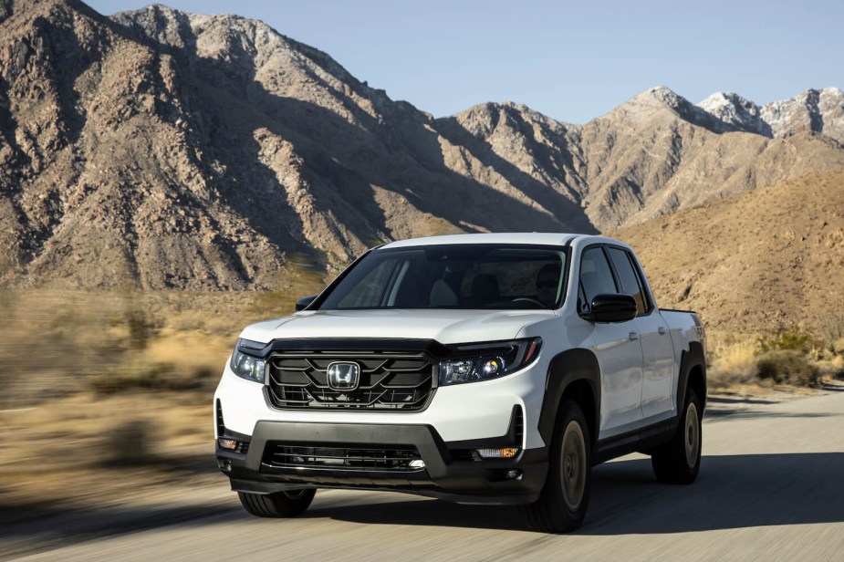 A white Honda Ridgeline truck drives through the mountains, a blue sky visible in the background.