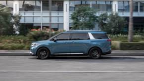 The side profile of a blue Kia Carnival driving on the street in front of a building.